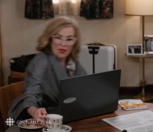 a woman sits at a table with a laptop and a cup of coffee with the hashtag schitts creek on the bottom