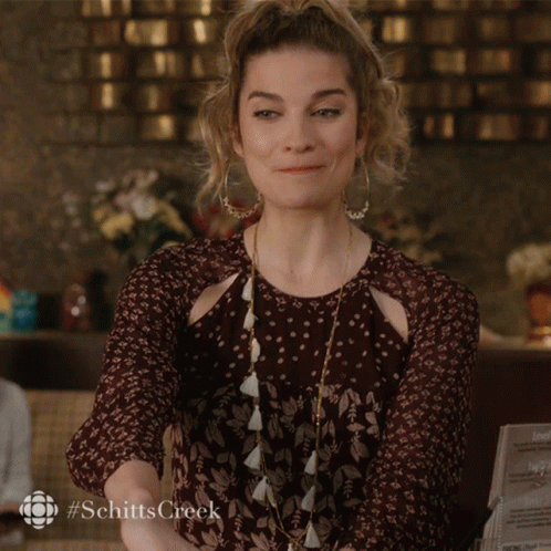 A Woman Sits At A Table With Her Hands On Her Chin While Holding A Cell Phone That Says #Schittscreek On It GIF