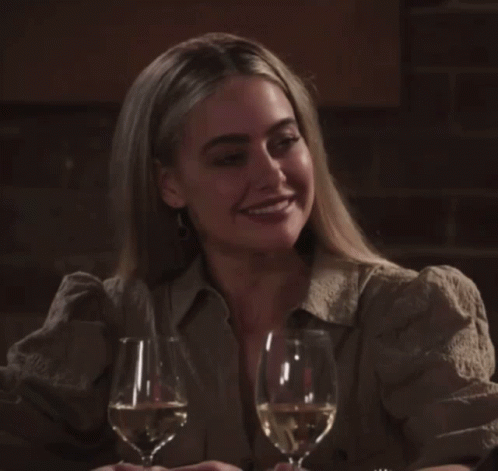 A Woman Sits At A Table With Two Glasses Of Wine In Front Of Her GIF