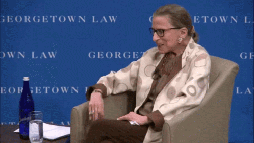 A Woman Sits In A Chair In Front Of A Georgetown Law Backdrop GIF