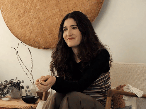 A Woman Sits In A Chair In Front Of A Wicker Wall Hanging GIF