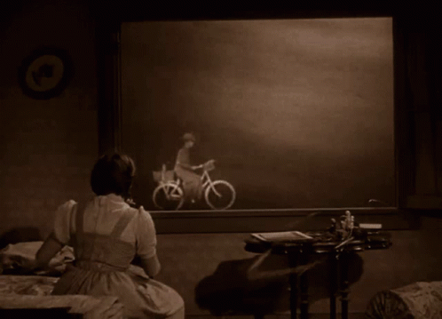 A Woman Sits In Front Of A Picture Of A Man Riding A Bicycle GIF