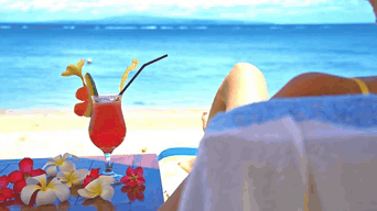 A Woman Sits On A Beach With A Drink On A Table GIF