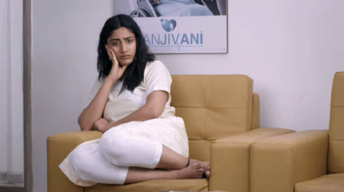 A Woman Sits On A Couch In Front Of A Poster That Says Anjivani GIF