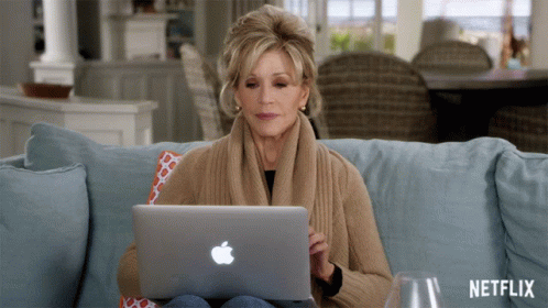 A Woman Sits On A Couch Using An Apple Laptop With Netflix Written On The Bottom GIF