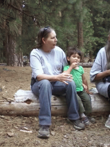 A Woman Sits On A Log Holding A Child Who Is Eating Something GIF