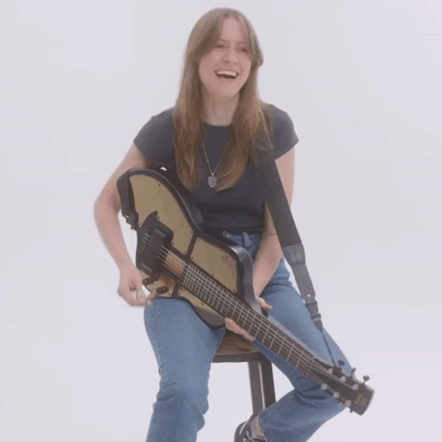 A Woman Sits On A Stool Holding A Guitar And Smiling GIF