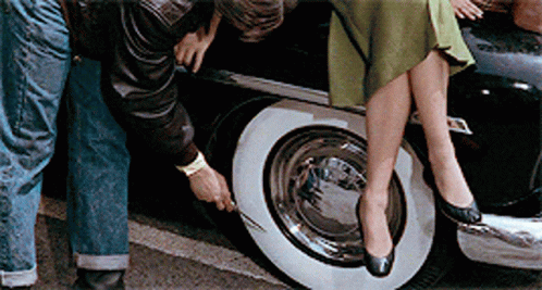 A Woman Sits On The Back Of A Car While A Man Checks The Tire GIF