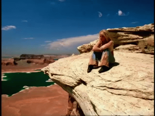A Woman Sits On The Edge Of A Rocky Cliff Overlooking A Lake GIF
