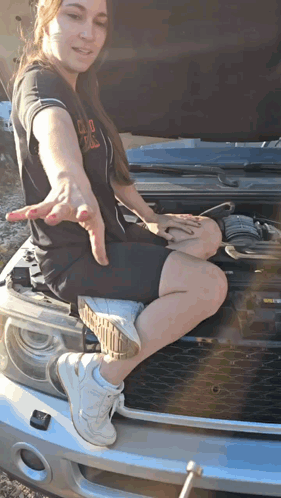 A Woman Sits On The Hood Of A Car With The Hood Open GIF