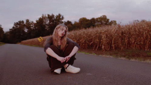 A Woman Sits On The Side Of A Road In Front Of A Cornfield GIF
