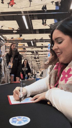 A Woman Sitting At A Table Signing A Piece Of Paper With A Pen GIF