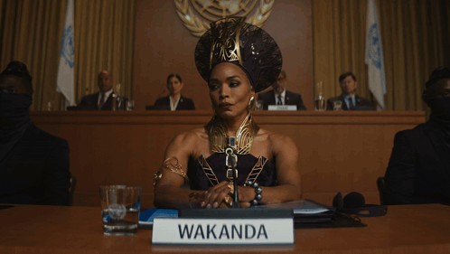 A Woman Sitting At A Table With A Sign That Says Wakanda On It GIF