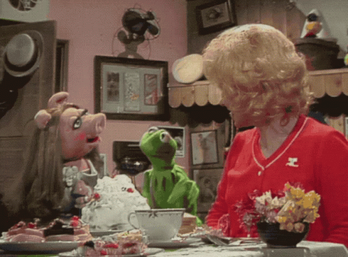 A Woman Sitting At A Table With Muppets Including Kermit GIF