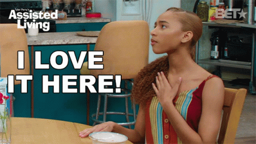 A Woman Sitting At A Table With The Words I Love It Here On The Screen GIF