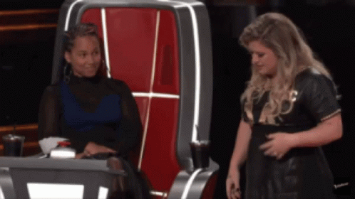A Woman Sitting In A Chair Talking To Another Woman In A Black Dress GIF