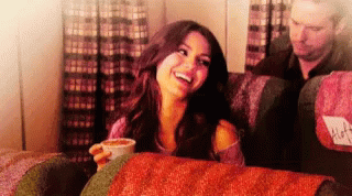 A Woman Sitting On A Couch With A Cup Of Coffee And A Man Behind Her With A Seat Cushion That Says Alfa GIF