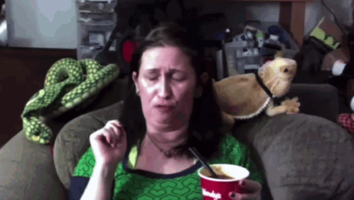 A Woman Sitting On A Couch With A Cup Of Ice Cream And A Spoon In Her Hand GIF