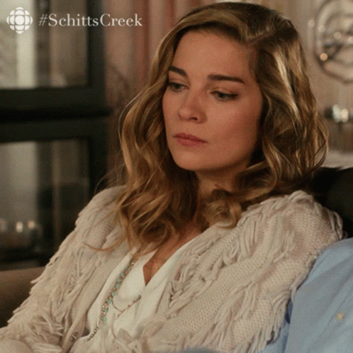 A Woman Sitting On A Couch With #Schitts Creek Written On The Bottom Right GIF