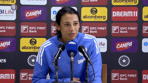 A Woman Speaking Into A Microphone In Front Of A Wall Of Advertisements For Campa And Atomberg GIF