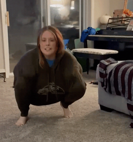 A Woman Squatting Down In A Living Room Wearing A Hoodie That Says Reebok GIF