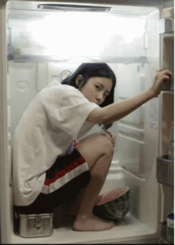 A Woman Squatting In An Open Refrigerator With A Watermelon In The Fridge GIF