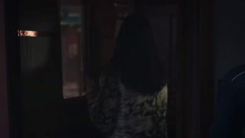 A Woman Standing In A Dark Room Looking Out A Window . GIF