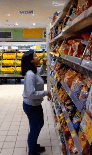 A Woman Standing In A Grocery Store Reaching For A Bag Of Chips GIF