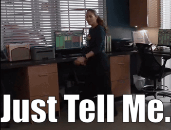 A Woman Standing In Front Of A Desk With The Words Just Tell Me Written Above Her GIF
