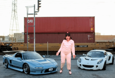 A Woman Stands Between Two Sports Cars In Front Of A Container That Says Cai GIF