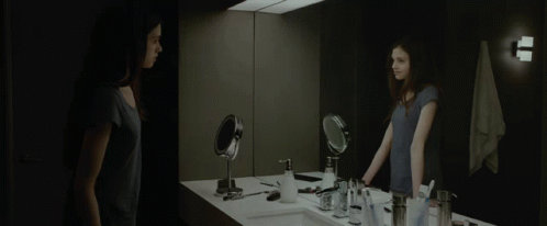 A Woman Stands In Front Of A Bathroom Mirror Looking At Herself GIF