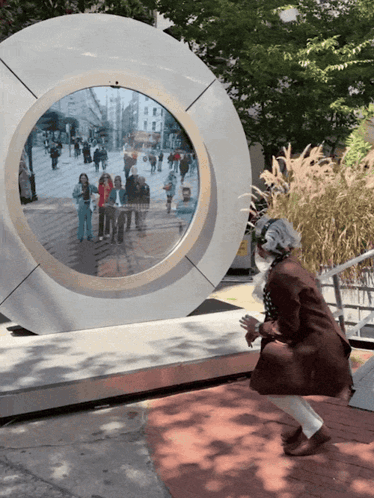 A Woman Stands In Front Of A Circular Sculpture With A Reflection Of People Walking Down A Street GIF