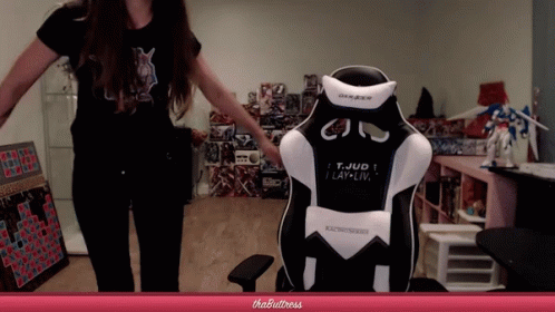 A Woman Stands In Front Of A Gaming Chair That Says T. Jud I Lay-Liv GIF