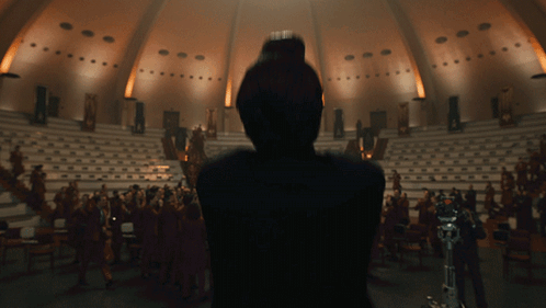 A Woman Stands In Front Of A Large Audience In A Dark Auditorium GIF