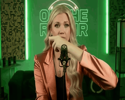 A Woman Stands In Front Of A Microphone In Front Of A Neon Sign That Says Othe R GIF