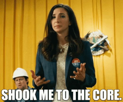 A Woman Stands In Front Of A Yellow Curtain With The Words Shook Me To The Core Above Her GIF
