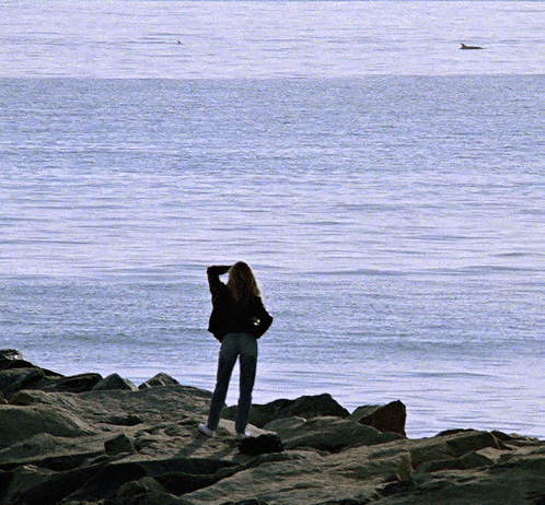 A Woman Stands On A Rocky Shore Looking Out Over The Ocean GIF