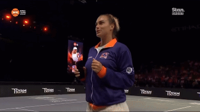 a woman stands on a tennis court wearing a blue and orange jacket