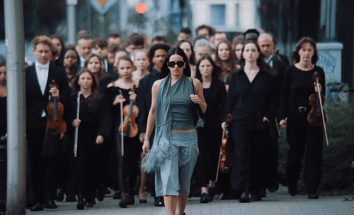 A Woman Walking In Front Of A Large Group Of People Holding Violins GIF
