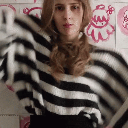 A Woman Wearing A Black And White Striped Sweater Is Standing In Front Of A Wall With Pink Drawings On It GIF