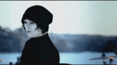 A Woman Wearing A Black Beanie And A Black Turtleneck Is Standing In Front Of A Body Of Water . GIF