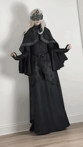 A Woman Wearing A Black Cape And A Mask Stands In Front Of A White Wall GIF