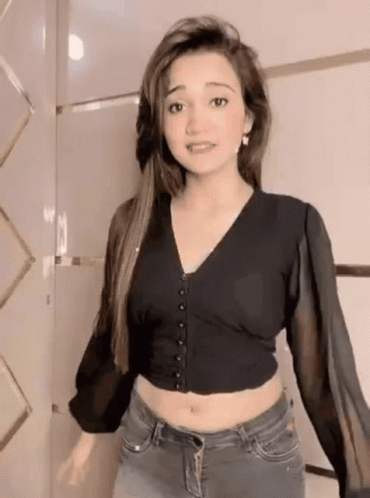 A Woman Wearing A Black Crop Top And Jeans Is Standing In Front Of A Wall . GIF
