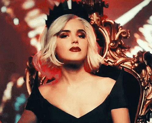 A Woman Wearing A Black Dress And A Crown Is Sitting On A Chair . GIF
