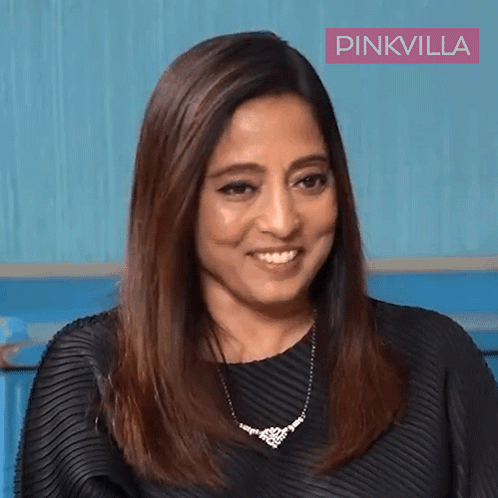 A Woman Wearing A Black Dress And A Necklace Is Smiling In Front Of A Pinkvilla Banner GIF