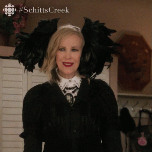 A Woman Wearing A Black Feathered Headband With The Word Schitts Creek Written On The Bottom GIF