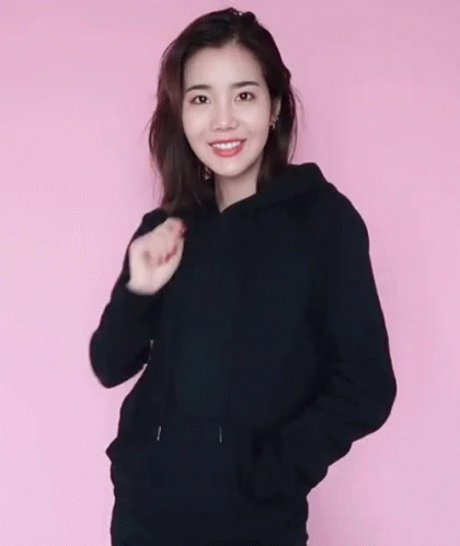A Woman Wearing A Black Hoodie And A Black Hat Against A Pink Background GIF