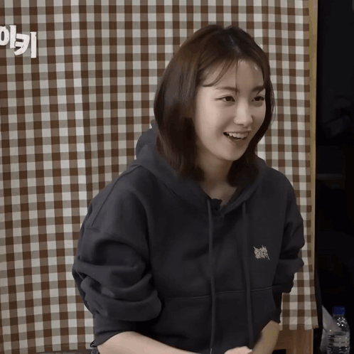 A Woman Wearing A Black Hoodie Smiles In Front Of A Checkered Tablecloth GIF