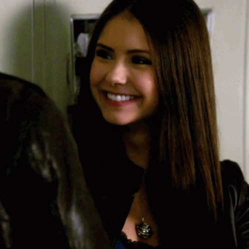 A Woman Wearing A Black Jacket And A Black Necklace Smiles GIF
