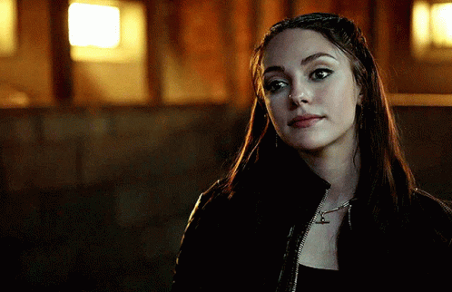 A Woman Wearing A Black Jacket And A Necklace With The Letter T On It GIF